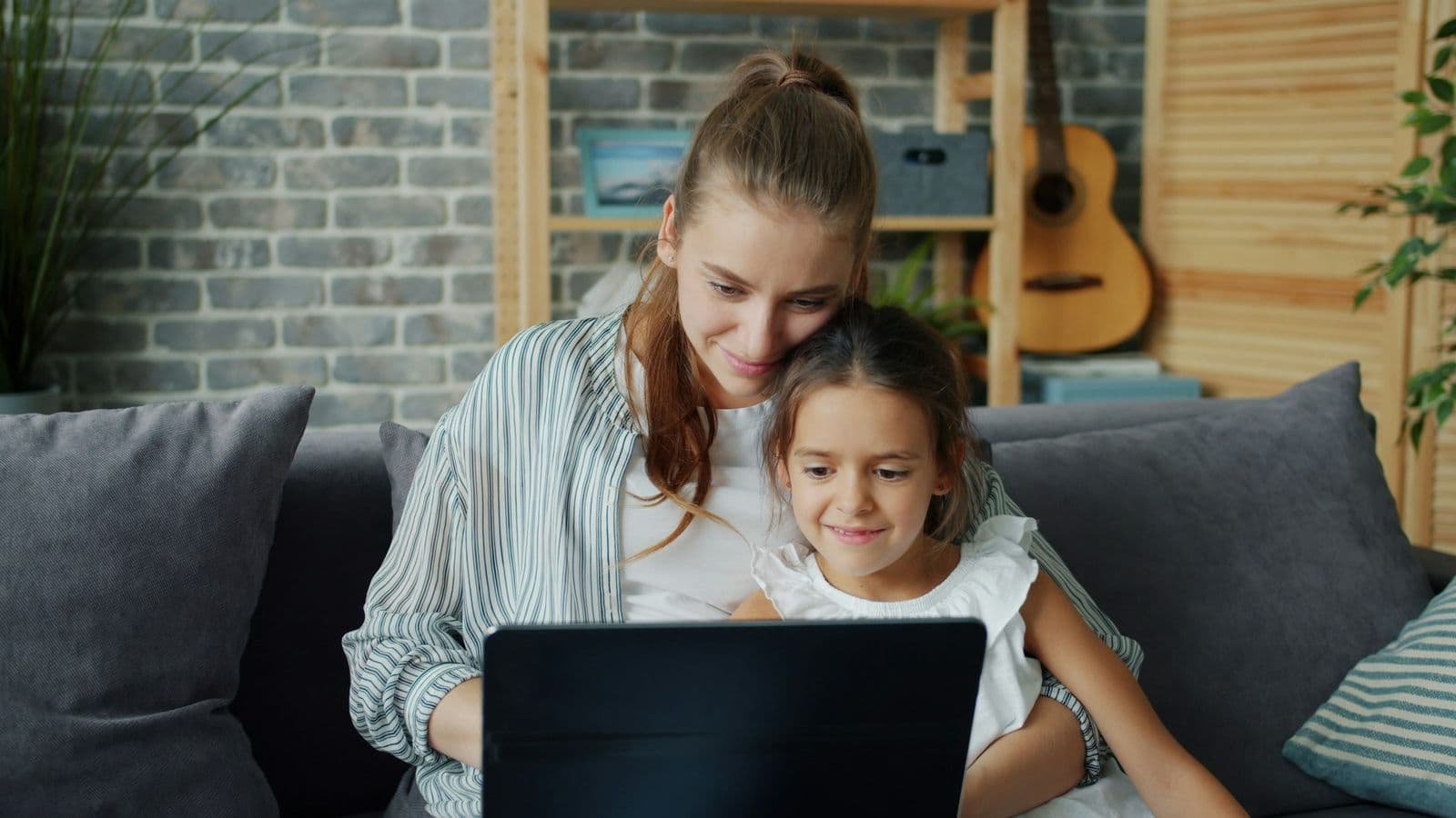 A mother and her young daughter smiling at a laptop on the couch at home